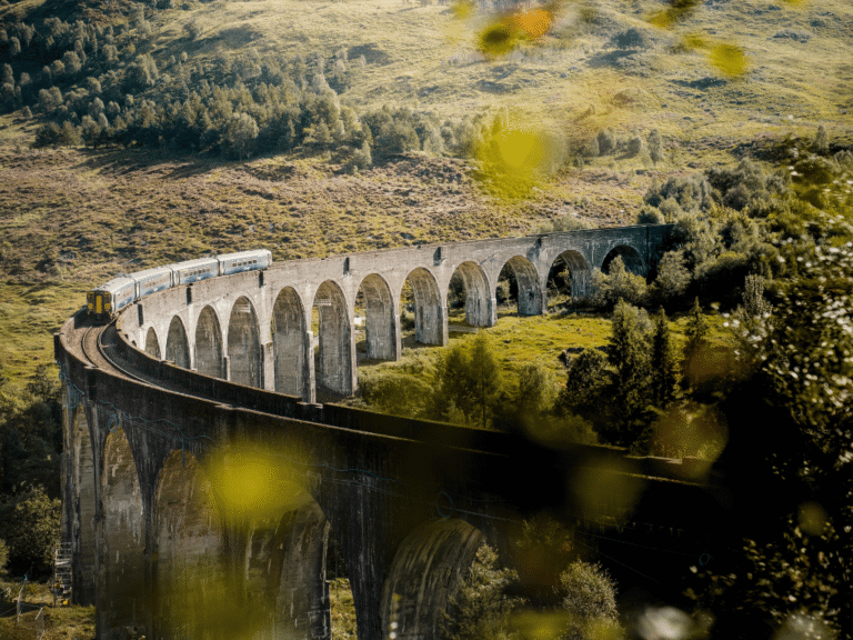 A passenger train crossing a large bridge in Europe, elevated above a river and surrounded by natural landscape, with hills and open sky in the background.