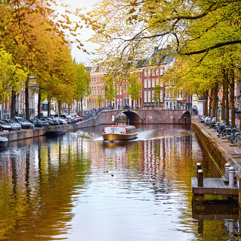 Historic canal in Amsterdam, Netherlands, lined with traditional narrow Dutch houses and trees reflected in the calm water, with small boats passing under a stone bridge in the city centre.