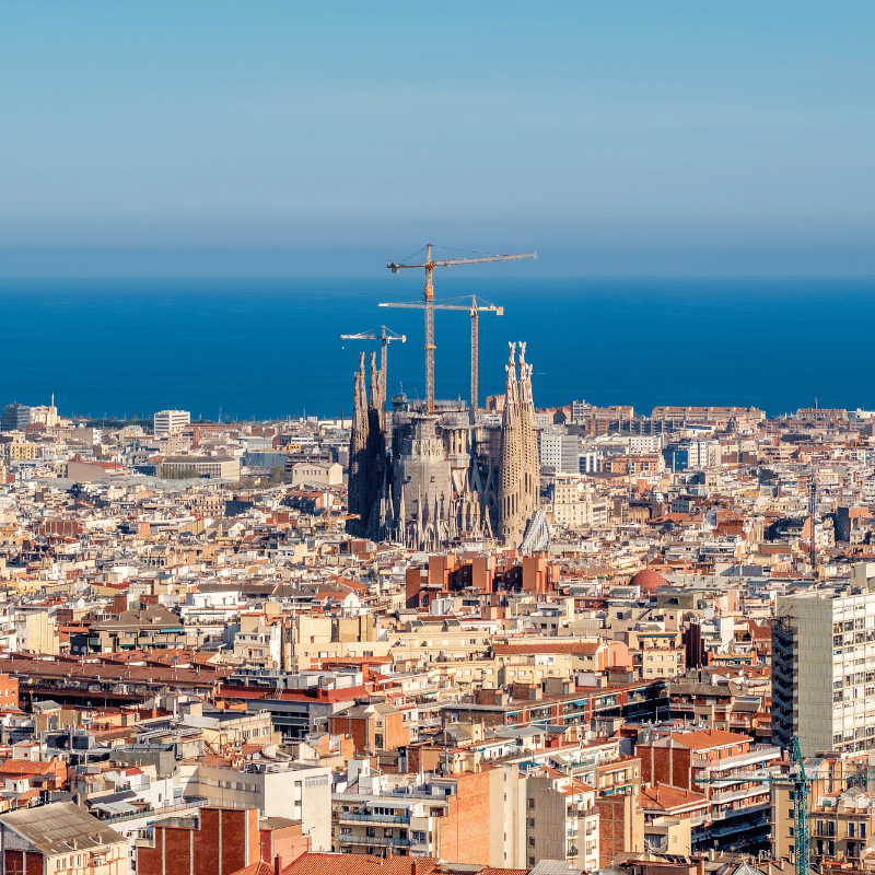 A panoramic view of Barcelona, Spain, featuring Mediterranean rooftops, modern and historic architecture, and the sea visible in the distance. The city skyline stretches under a bright blue sky, blending urban life with coastal scenery.
