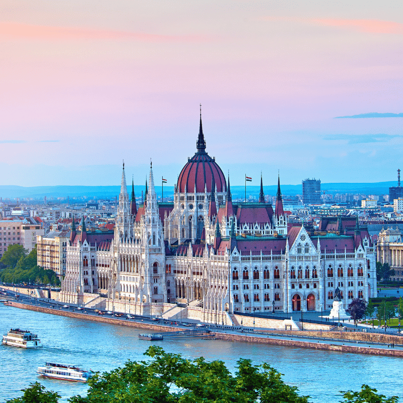Panoramic view of Budapest, Hungary, featuring the Hungarian Parliament Building along the Danube River, with historic architecture and bridges connecting Buda and Pest under a clear sky.