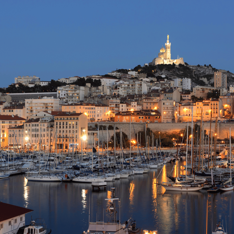 View of Marseille, France, featuring the Old Port (Vieux-Port) with colourful boats docked along the harbour, Mediterranean waters, and historic buildings under a bright blue sky.
