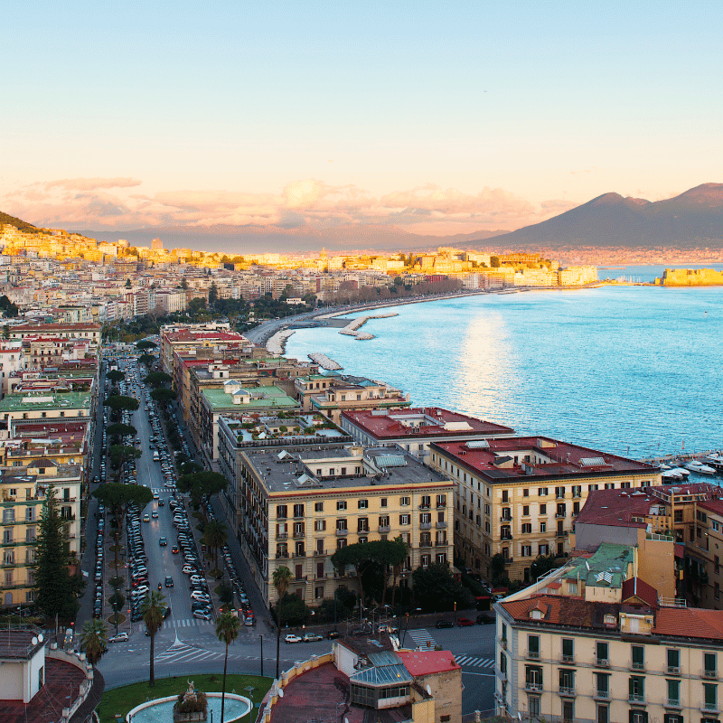 Panoramic view of Naples, Italy, featuring the city’s waterfront along the Bay of Naples with Mount Vesuvius rising in the background under a clear Mediterranean sky.