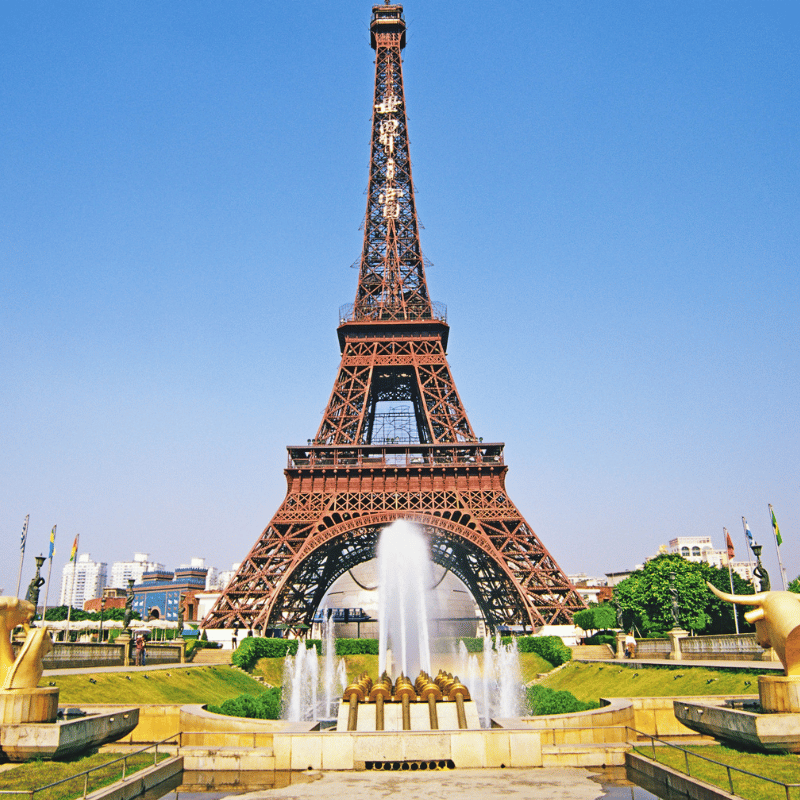 The Eiffel Tower in Paris, France, a tall iron lattice structure rising above the city skyline. The monument stands prominently against the sky, surrounded by green gardens and classic Parisian architecture.