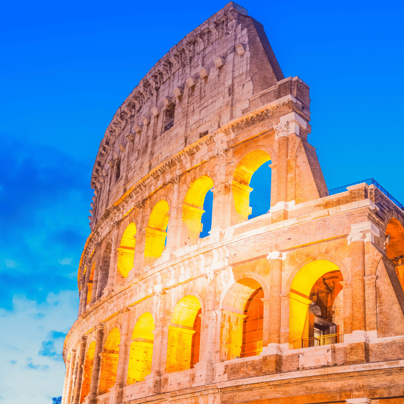 A panoramic view of Rome, Italy, featuring historic buildings, terracotta rooftops, and iconic domes rising above the city skyline. The scene includes narrow streets and classical architecture, surrounded by Mediterranean trees under a clear blue sky.