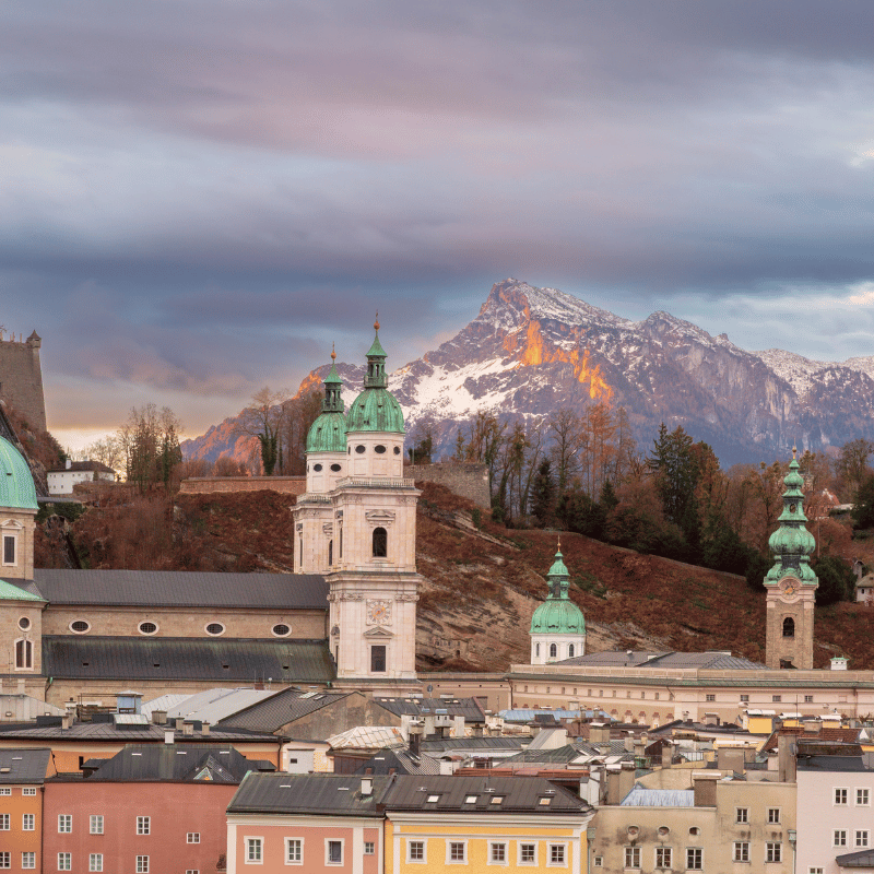 View of Salzburg, Austria, showing the historic Old Town with baroque architecture and Hohensalzburg Fortress overlooking the city, surrounded by alpine scenery under a clear sky.