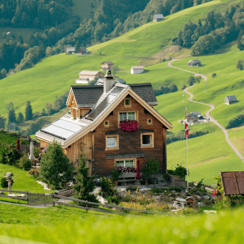 A small traditional house in the Swiss countryside, surrounded by green meadows and alpine mountains under a clear blue sky.