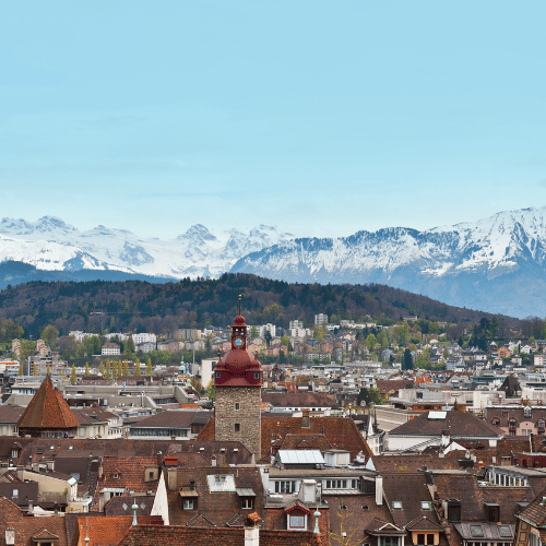View of Lucerne, Switzerland, featuring the historic Chapel Bridge crossing the Reuss River, colourful old town buildings, and alpine mountains in the background.