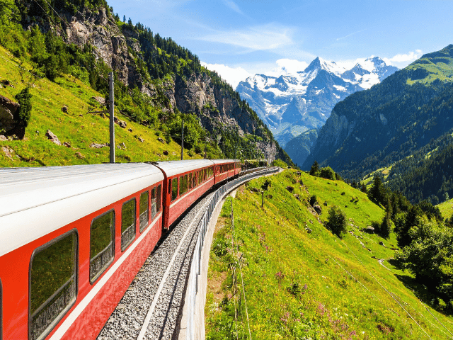 A passenger train travelling through the Swiss Alps, surrounded by green valleys, alpine peaks, and clear blue skies, with panoramic mountain scenery visible along the route.