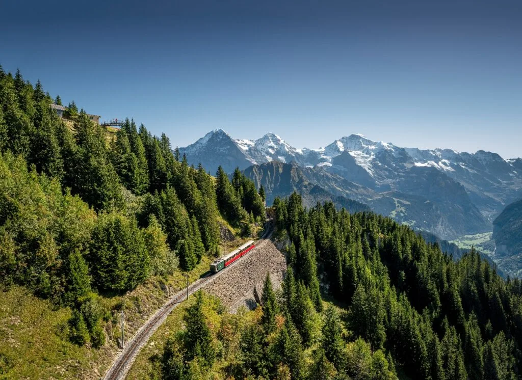 Red mountain train climbing through pine forest towards the snow-capped Eiger, Mönch and Jungfrau